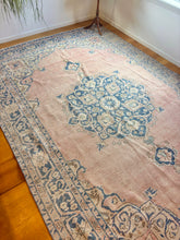 Load image into Gallery viewer, A rectangular vintage-style area rug with a faded pink field, blue medallion, and beige floral border, shown on a wooden floor in a room corner.